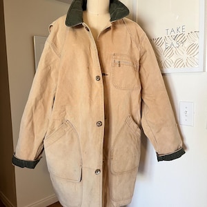 Può includere: Una giacca di tela beige con colletto e polsini in velluto a coste verde. La giacca ha una chiusura con bottoni, due grandi tasche sul davanti e una tasca con zip sul lato sinistro.