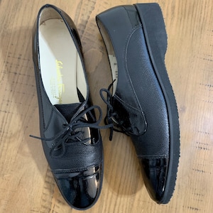 Pu&ograve; includere: Un paio di scarpe stringate in pelle nera con punta in vernice. Le scarpe hanno una suola in gomma spessa e sono realizzate da Salvatore Ferragamo.