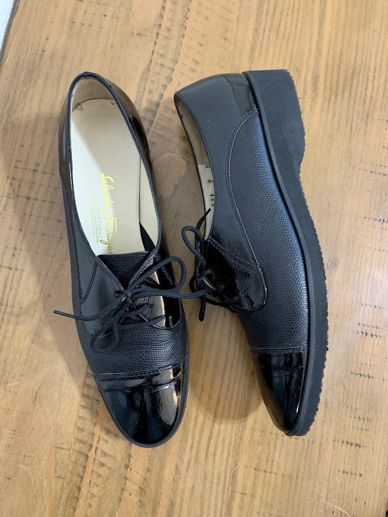 Pu&ograve; includere: Un paio di scarpe stringate in pelle nera con punta in vernice. Le scarpe hanno una suola in gomma spessa e sono realizzate da Salvatore Ferragamo.