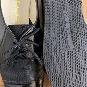 Pu&ograve; includere: Un paio di scarpe stringate in pelle nera con punta in vernice. Le scarpe hanno una suola in gomma con un motivo a griglia e il testo "Salvatore Ferragamo" sulla suola.