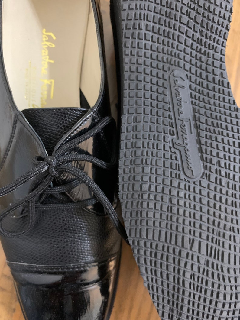 Pu&ograve; includere: Un paio di scarpe stringate in pelle nera con punta in vernice. Le scarpe hanno una suola in gomma con un motivo a griglia e il testo "Salvatore Ferragamo" sulla suola.