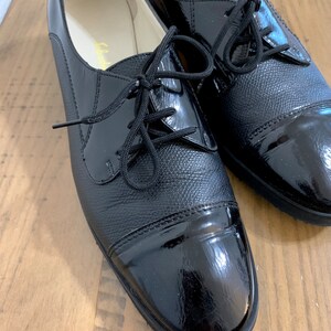 Pu&ograve; includere: Un paio di scarpe stringate in pelle nera con punta in vernice. Le scarpe hanno una punta arrotondata e un tacco basso.