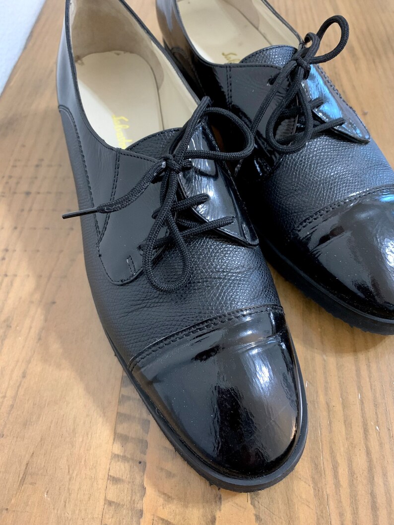 Pu&ograve; includere: Un paio di scarpe stringate in pelle nera con punta in vernice. Le scarpe hanno una punta arrotondata e un tacco basso.