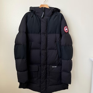 Puede incluir: Parka de invierno negra con capucha, colgada de una percha de madera. La chaqueta tiene un diseño acolchado, dos bolsillos delanteros y un logotipo circular rojo y blanco en la manga izquierda. La parka está hecha de material resistente al agua.