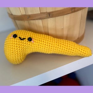 May include: A handmade, yellow crochet pancreas with black eyes and a smiling mouth. The plush organ is resting on a white shelf, with a wooden basket in the background. The pancreas is a whimsical, educational item.