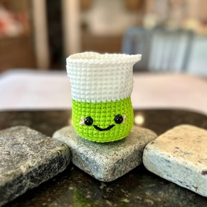 Può includere: Una piccola tazza all'uncinetto verde e bianca con un viso sorridente. La tazza è appoggiata su una pietra grigia.
