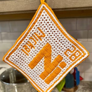 May include: Crocheted orange and white pot holder with the word "NICK" and the letter "N" in orange. The pot holder has a diamond shape with a loop for hanging.