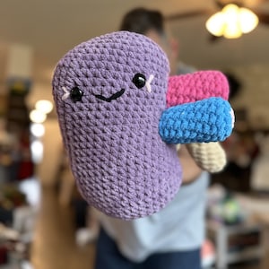 May include: A handmade, plush, purple kidney-shaped toy with a smiling face, black eyes, and pink accents. Attached are pink, blue, and beige crocheted sections. The toy is soft and cuddly, perfect for children or as a unique gift.