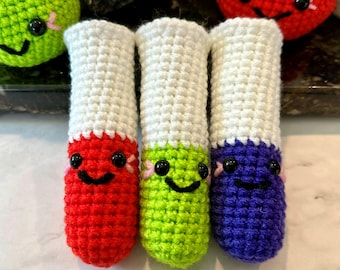 May include: Three handmade crochet markers in red, green, and purple, each with a white top. The markers have smiling faces with black eyes and pink accents. Additional crochet items are visible in the background.