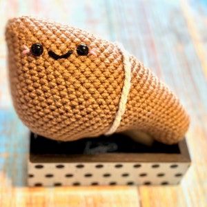 May include: A crocheted brown plush toy shaped like a human liver with a smiling face and black eyes. The toy is sitting on a black and white polka dot box.