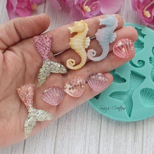 Könnte beinhalten: Eine türkisfarbene Silikonform mit Vertiefungen für Seepferdchen, Meerjungfrauenschwänze und Muscheln, zusammen mit losen Harzanhängern in passenden Formen. Die Anhänger umfassen glitzernde rosa und silberne Meerjungfrauenschwänze, gelbe und blaue Seepferdchen sowie durchscheinende rosa Muscheln mit Goldflocken. Die Form ist mit dem Text Sissy's Creations geprägt.