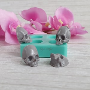 May include: Four gray skull-shaped silicone molds on a turquoise silicone sheet. The molds are arranged in a square pattern. The sheet has the text "Sugga Crafts" printed on it.