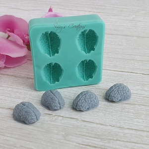 Puede incluir: Un molde de silicona con cuatro cavidades en forma de cerebro. El molde es verde turquesa y tiene el texto "Sugars Crafts" impreso en él. Hay cinco objetos grises en forma de cerebro al lado del molde.