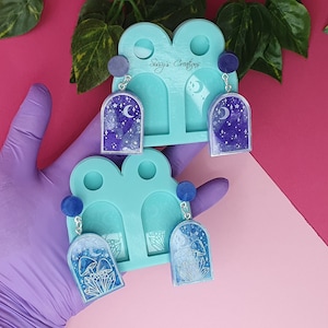 Puede incluir: Dos pares de pendientes azules y plateados con un diseño de luna y estrellas. Los pendientes se muestran en un molde de silicona azul con el texto "Sugary Crafters".