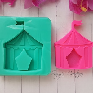 Könnte beinhalten: Eine grüne Silikonform und eine rosa Plastikform eines Zirkuszeltes mit einer sternförmigen Öffnung. Die Formen befinden sich auf einer weißen Holzoberfläche.