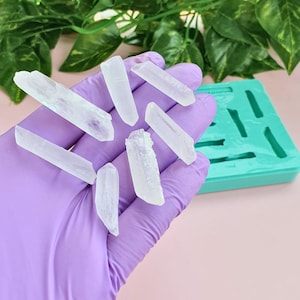 Op de afbeelding: Verschillende heldere, doorschijnende, grillige kristalpunten worden vastgehouden in een hand met een paarse nitrilhandschoen. Een rechthoekige, groenblauwe siliconen mal met meerdere uitsparingen in de vorm van kristalpunten ligt ernaast.