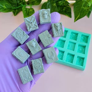 Puede incluir: Una colección de diez pequeños dijes de resina gris claro con forma de libro que presentan varios diseños en relieve, incluyendo una calavera, un pentagrama, un ojo y una luna creciente, junto a un molde de silicona turquesa con nueve cavidades.