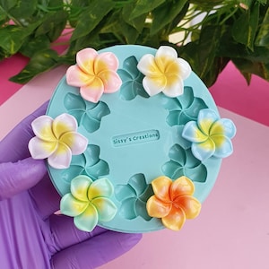 May include: A round, light blue silicone mold featuring multiple flower-shaped cavities and the embossed text Sissy's Creations. Five finished plumeria flower embellishments in pink, white, blue, green, and orange are placed on the mold surface.