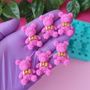 Puede incluir: Seis encantos de oso rosa de rosa con lazos dorados. Los encantos están hechos de arcilla polimérica y tienen aproximadamente 5 cm de alto. Los encantos se muestran en un guante morado y un molde de silicona azul.