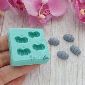 May include: A light teal silicone mold with four pumpkin shapes and the text "Sissy's Creations". Four small, gray pumpkin-shaped soaps are next to the mold. Pink orchids are in the background.