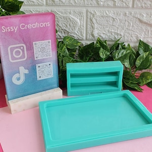 May include: A teal-coloured rectangular tray, a small teal shelf with two levels, and a pink and blue gradient stand with the text "Sissy Creations" and social media icons. Green leafy plants and a white brick wall are in the background.