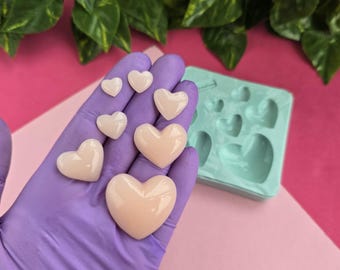 Puede incluir: Un molde de silicona con múltiples cavidades en forma de corazón, que se muestra con varias piezas de resina en forma de corazón de diferentes tamaños, que van de pequeñas a grandes, en un color rosa pálido.