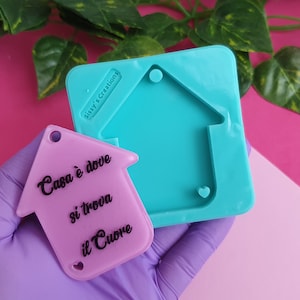 Puede incluir: Un blanco de resina de color morado claro en forma de casa con el texto "Casa è dove si trova il Cuore" en letras negras. El blanco está siendo sostenido por una mano enguantada. También se puede ver un molde de silicona turquesa en forma de casa en la imagen.