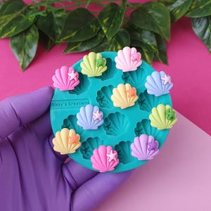 Puede incluir: Un molde de silicona turquesa lleno de adornos coloridos en forma de concha. Las conchas son en tonos rosa, amarillo, azul y verde, cada una adornada con una pequeña estrella de mar. El molde tiene el texto "Sissy's Creations".