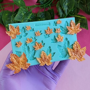 Puede incluir: Un molde de silicona turquesa con impresiones de hojas y varias decoraciones de hojas de arce doradas. El molde tiene el texto "Sissy's Creations". Las hojas son de varios tamaños y están dispuestas sobre un fondo rosa y verde.