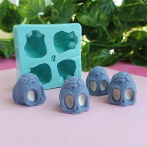 May include: A light blue silicone mold with four skull shapes and the letter "S" is next to several small, blue skull-shaped candies with white eye sockets and brain details. The candies are on a pink surface.