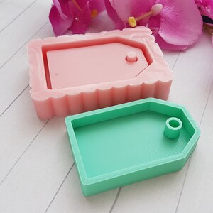 Puede incluir: Dos moldes de silicona para hacer etiquetas de resina. El molde de la izquierda es rosa y tiene un borde festoneado. El molde de la derecha es verde y tiene un borde liso. Ambos moldes tienen un agujero para una cuerda o cinta.