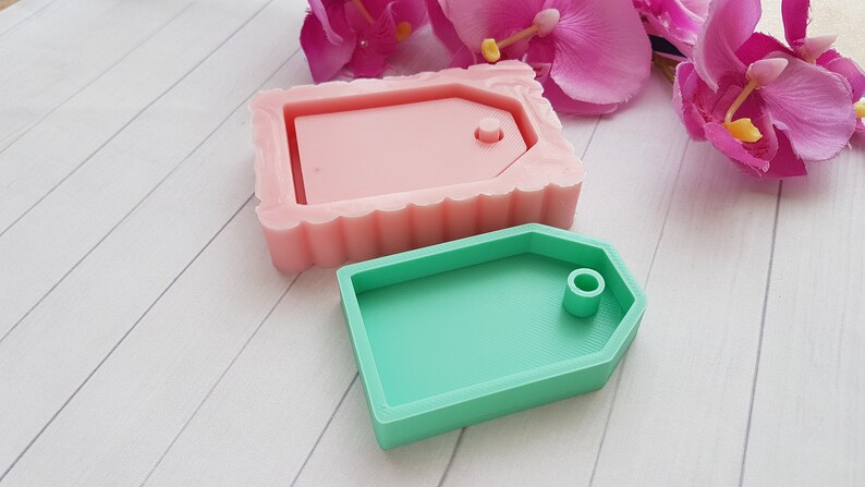 Puede incluir: Dos moldes de silicona para hacer etiquetas de resina. El molde de la izquierda es rosa y tiene un borde festoneado. El molde de la derecha es verde y tiene un borde liso. Ambos moldes tienen un agujero para una cuerda o cinta.