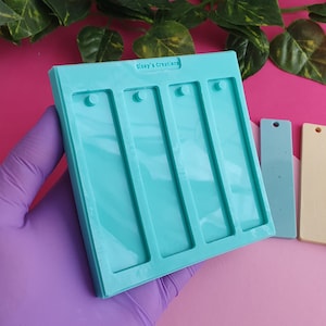 Peut inclure: Un moule en silicone turquoise avec quatre cavités rectangulaires pour la fabrication de porte-clés en résine. Le moule comporte de petits cercles en haut de chaque cavité pour ajouter un anneau de saut. Trois porte-clés en résine finis sont présentés à côté du moule, un bleu, un jaune et un avec des paillettes et le texte "Ti Amo Papa".