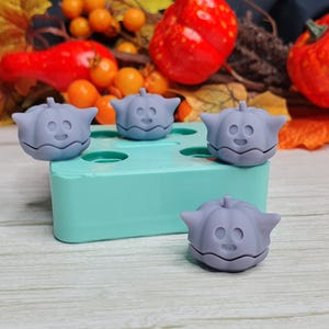 May include: Four purple silicone molds shaped like pumpkins with a cute face on each. The molds are sitting on a light blue silicone tray.