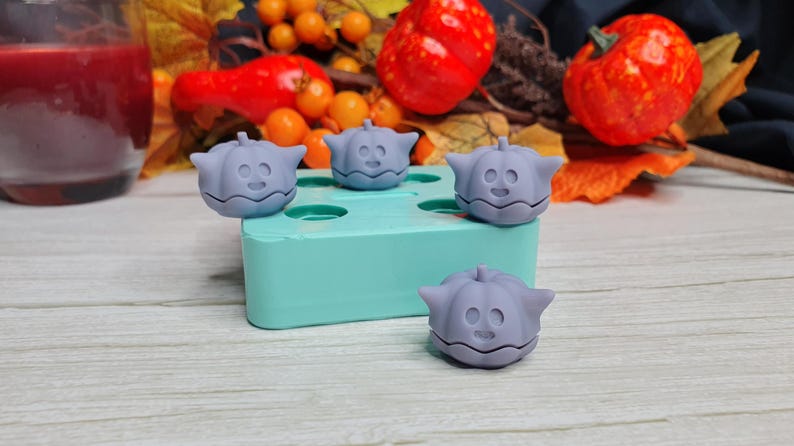 May include: Four purple silicone molds shaped like pumpkins with a cute face on each. The molds are sitting on a light blue silicone tray.