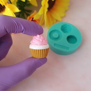 Puede incluir: Un cupcake rosa y blanco con glaseado marrón se sostiene en una mano enguantada de morado. Un molde de silicona turquesa con tres formas de cupcake está en el fondo.