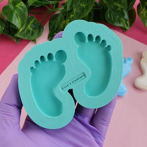 Peut inclure: Un moule en silicone bleu turquoise en forme de deux empreintes de pieds de bébé. Le moule est tenu dans une main gantée. Le texte "Sissy's Creations" est imprimé sur le moule.