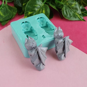 May include: A teal silicone mold with two gray bat-shaped castings. The mold is on a pink surface.