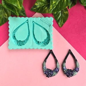 Puede incluir: Pendientes en forma de lágrima con diseño floral, con marco negro y rosas moradas. También se ve un molde de silicona turquesa con la misma forma de pendiente, con el texto "Sissy's Creations". Los pendientes se muestran sobre un fondo rosa.