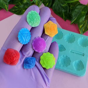 Peut inclure: Sept symboles de chakra colorés, chacun avec un design et une couleur uniques, sont affichés sur une main gantée de violet. Les symboles sont en résine et sont disposés en arc-en-ciel. Un moule en silicone avec sept symboles de chakra est également présenté sur l'image.