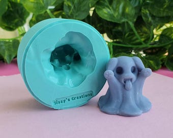 May include: A light blue silicone mold with the text "Sissy's Creations" next to a small, gray ghost dog figurine. The ghost dog has its tongue sticking out and its arms raised. The mold is round and has a cavity in the shape of the ghost dog.