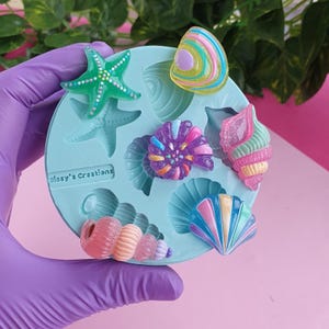 Pode incluir: Um molde de silicone circular azul-claro com várias cavidades com tema marinho, incluindo uma estrela-do-mar, conchas em espiral e amêijoas. Várias peças de resina coloridas e brilhantes estão colocadas no molde, que tem gravado o texto Sissy's Creations.
