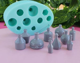 Op de afbeelding: Een lichtblauwe siliconen mal met diverse ronde en geometrische holtes, vergezeld van tien kleine, gladde, grijze miniatuurflesjes in verschillende vormen en maten, gerangschikt voor de mal op een vlakke ondergrond.