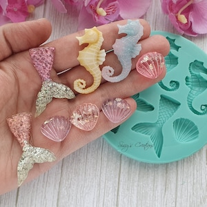 以下が含まれることがあります： シーホース、貝殻、人魚の尻尾など、さまざまな海のテーマの形状を持つシリコンモールド。モールドは青色で、「Sugars Crafts」というテキストが書かれています。画像には、ピンク、イエロー、ブルーのシーホース、ピンクの貝殻、ピンクとシルバーの人魚の尻尾など、完成したレジン作品もいくつかあります。