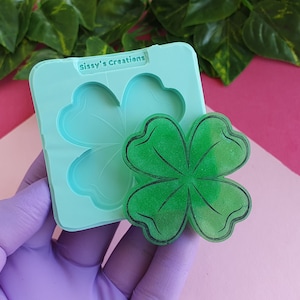 Può includere: Una fusione in resina di trifoglio a quattro foglie verde con uno stampo in silicone sullo sfondo. Lo stampo è etichettato "Sissy's Creations".