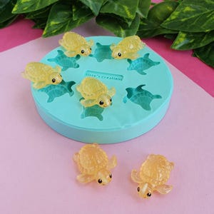 Könnte beinhalten: Eine hellblaue Silikonform mit dem Text "Sissy's Creations" enthält mehrere kleine, gelbe, schildkrötenförmige Kunstharzgüsse. Zwei weitere Schildkröten sind darunter platziert. Die Schildkröten haben sichtbare Augen und Flossen.