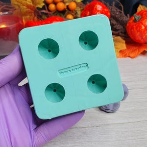 May include: A teal blue silicone mold with four round cavities. The mold is designed to create small, round objects. The mold has the text "Shay's Creations" printed on it.