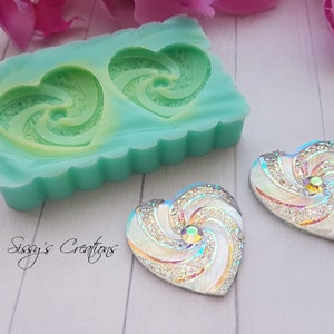 May include: A teal silicone mold with two heart-shaped cavities, each featuring a swirling design. Two finished heart-shaped embellishments with iridescent glitter and a central crystal are also present. The text "Sissy's Creations" is visible.