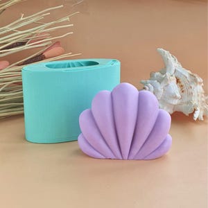 Può includere: Uno stampo in silicone azzurro per colate e un oggetto finito a forma di conchiglia color viola lavanda. Lo stampo è di forma ovale con un centro cavo, e l'oggetto a forma di conchiglia presenta creste verticali e una base arrotondata.
