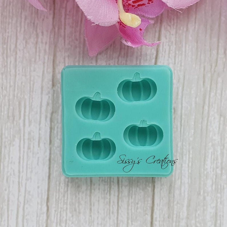 May include: A teal silicone mold featuring four pumpkin shapes. The mold has the text "Sissy's Creations" in cursive. The background includes a pink flower and a light wood-grain surface.